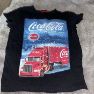 Svart Coca-Cola t-shirt med jultryck - Svart t-shirt från Coca-Cola med färgglatt tryck av den klassiska röda Coca-Cola-lastbilen i ett vintrigt snölandskap. Stor logga framtill och texten 'Holidays Are Coming'. Rund halsringning med röd detalj på insidan. Perfekt för dig som älskar retro och julkänsla.