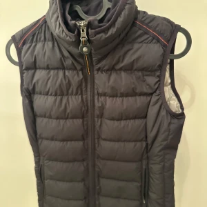 Svart dunväst Parajumpers XS - Snygg svart dunväst från Parajumpers i superlätt material. Västen har hög krage, dragkedja framtill och diskreta röda detaljer vid axlarna. Perfekt för lager-på-lager och har en sportig vibe med quiltad design och praktiska fickor.