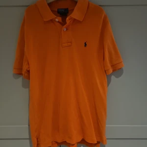 Orange pikétröja från Ralph Lauren - Snygg orange pikétröja från Polo Ralph Lauren med klassisk krage och två knappar framtill. Tröjan har korta ärmar och den ikoniska broderade loggan i mörkblått på bröstet. Perfekt för dig som gillar färg och stilren design.
