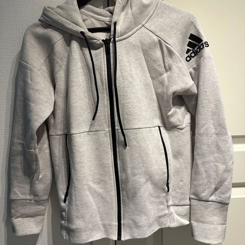 Grå kofta från adidas.  Adidas märke på vänster överarm.  Svarta detaljer och dragkedja.  Rymlig luva. . Hoodies.