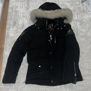 Moose Knuckles Jacka - En svar Mosse Knuckles jacka med vit päls för herr. En kort modell. Den är i nyskick knappt använd väldigt varm och stilig.