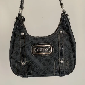 Guess väska - Väska av Guess. Det finns lite slitningar på kanten av axelremmen (se sista bilden) men annars jättefint skick. Bredd: 32 cm Höjd: 20 cm Längd av axelrem: 46 cm