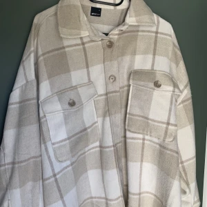 Skjortjacka  - Skön rutig skjortjacka i beige och vitt med två rymliga bröstfickor. Lös i modellen. Aldrig använd. Nytvättad såklart. 