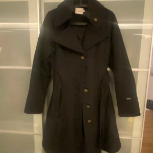 Vinterkappa figursydd - Figursydd kappa med stora bronsfärgade knappar och skärp i midjan. Saknas en knapp mitt på men går att flytta från kragen. Lång, slutar strax under knäna. Mörkgrå. 