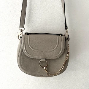 Axelremsväska med kedja - Fin axelremsväska i en varmgrå nyans med kedja. Justerbart band och gott om utrymme för att vara en liten väska 🌼