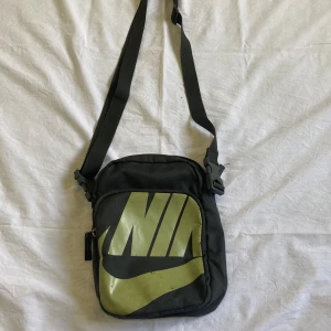Nike axelväska  - Nike axelväska med snöre man kan ta på och av enkelt och som man kan ändra längd på enkelt, den har ett stort fack med ett litet fack i och ett medelstort ytter fack. Helt ok skick lite sliten på märket men annars som helt ny