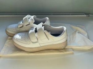 ACNE STUDIOSPerey Friend Sneakers - Vit, kalvskinn, perforerad detalj, logotyptryck i sidan, präglad logotyp i bak, rund tå, kardborreband i fram, innersula med logotyp, platt gummisula.