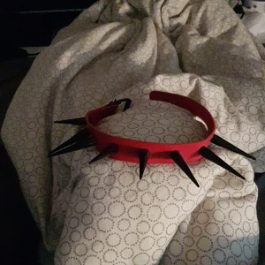Röd Choker - Röd choker med stora svarta spikar. Spikarna är inte vassa alls. Haft på mig den typ 8 gånger men inte så länge. Håller upp spikarna jätte bra och ser jätte bra ut på alla hals formar. 