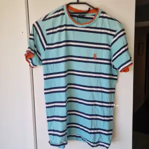 Ljusblå t-shirt med vit-blå ränder och orangea detaljer. Storlek XS. Bra skick, kanppt använd.