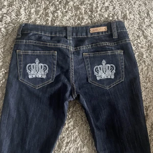 Lågmidjade jeans med kronor - Jeans som liknar victoria beckham, mörkblåa utsvängda med kronor på bakfickorna. Midjemått- 74cm, innerbenslängd- 81cm. Jag är 1,71