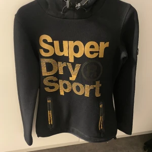 Superdry huvtröja  - Svart Superdry huvtröja med guldigt tryck.