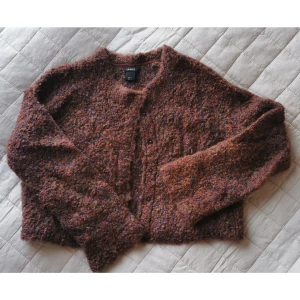 Höstig kofta i ullmix - Höstig stickad kofta med tryckknappar. Kort modell med något utsvängd ärm. Ullblandning. Melerad i kastanjebrun och blå. LINDEX strl S. Axelbredd ca 40 cm, längd ca 45 cm. 