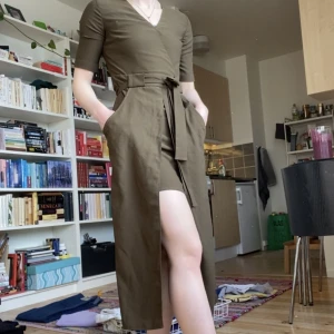 Klänning - Militär-/olivgrön klänning med slits framtill. Dragkedja i sidan och bälte för snygg passform, kan knytas antingen fram eller baktill. Går till mitten av vaderna på mig som är 160cm.
