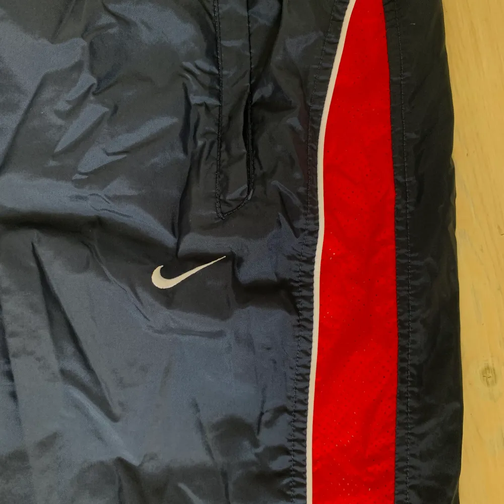 Nike trackpants/prasselbyxor i mörkblå med röda detaljer! Mycket bra skick! Dragkedjor i fickorna! Stl L men passar även M!. Jeans & Byxor.