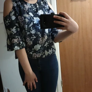 Svart blus med blommor - Med hål på axlarna, använd 3 gånger, kommer i jätte fint skick, tvättas också innan leverans, kostar 150 kr egentligen, Perfekt för fester