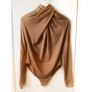  - Kamelfärgad body från H&M. Hög i halsen med drapering fram och dragkedja bak. Aldrig använd och prislappen sitter kvar. Pris exklusive frakt. 