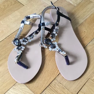  - Superfina sandaler med stenar, köpta i Köpenhamn. Använts enbart en gång pga för stora. Strl 38 (jag har 37). Se tredje bilden där det fattas en sten, ej märkbart eftersom resten är guldfärgat. Passar lika snyggt till vardags som till fest! Säljer ett par likadana med rosa stenar i Strl 37 