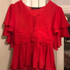  - Röd blus i chiffong med volang från Zara, storlek S (passar XS/S) dekorativ "snörning" i ringningen och åtdragen med drapering i midjan, dold dragkedja i ena sidan men även öppen nedtill på båda sidorna som ett sprund Fint skick!!! Använd fåtal gånger 