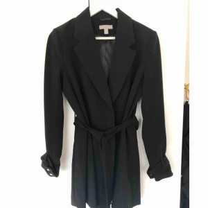 Svart kappa - Helt ny oanvänd kappa från HM i strl 38. Dubbla tryck knappar i midjan samt bälte. Guld knapp detaljer i ärmarna & axelvaddar. Frakt tillkommer på 79kr