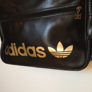  - Adidas airliner retroväska, svart med logga i guld. Snygg skol/arbetsväska med dragkedja i öppningen, ytterfack med dragkedja och innerfack samt mobilficka. Bra skick! Möts i Sthlm/frakt. 