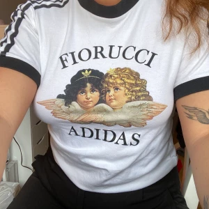 Fiorucci x Adidas T-Shirt  - Helt oanvänd Fiorucci x Adidas t-Shirt! 🤍🖤 Endast provad men lappen är tyvärr bortklippt då jag trodde jag skulle använda den... Storlek UK12 men ganska liten så skulle säga UK10. Nypris 499kr, budgivning från 150kr + frakt!! 🤩👍🏼