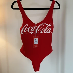 Body CocaCola - Röd body med CocaCola-tryck. Figursydd och stretchigt material. Aldrig använd, prislapp kvar, nypris 199 kr.