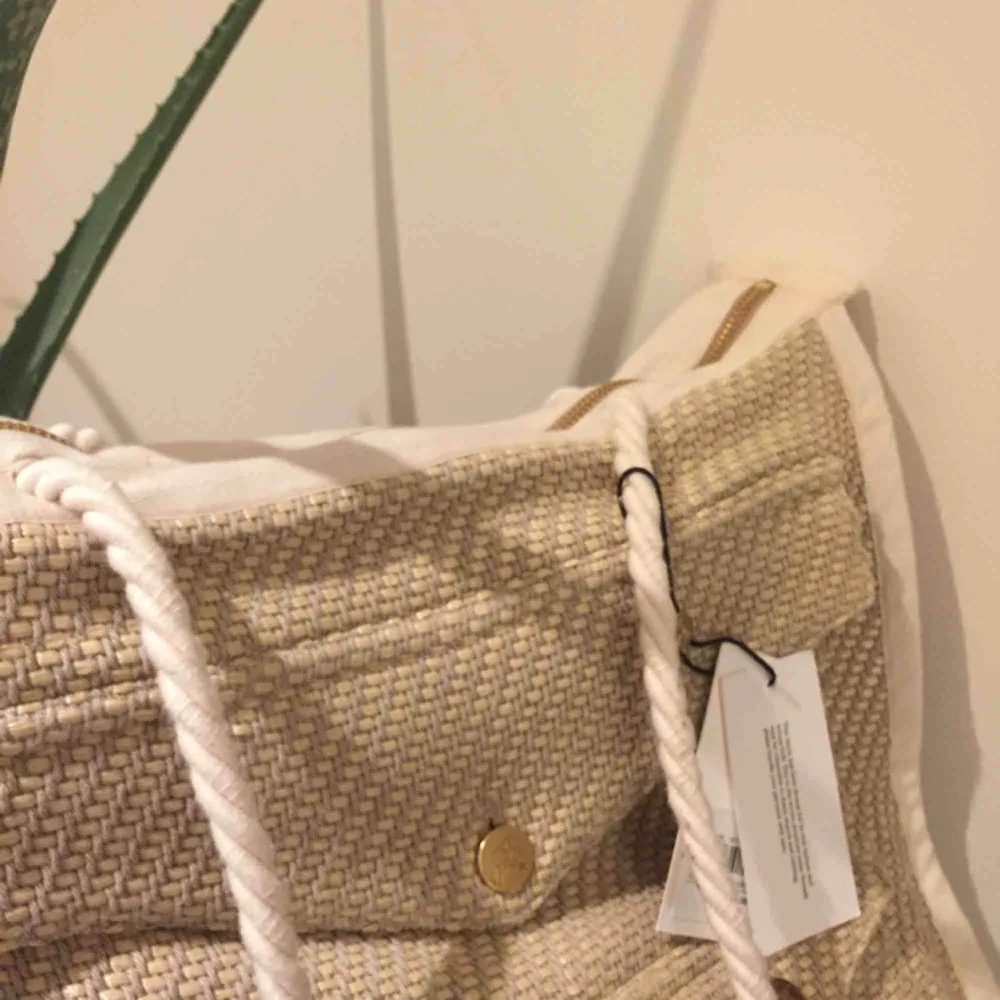 En helt ny väska från rodebjer, med alla prislappar kvar.   Modellen heter straw tote bag, superfint ljust tyg med guldiga o vita detaljer.   Nypris 1799:-. Väskor.