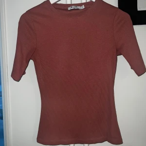 Rostfärgad ribbad topp från NA-KD - Säljer en rostfärgad ribbad topp från NA-KD med rund halsringning och korta ärmar. Toppen har en tight passform och är tillverkad i ett mjukt, stretchigt material som sitter snyggt på kroppen.