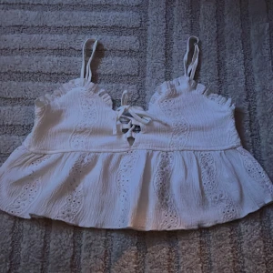 Vit broderad linnetopp med knyt - Söt vit linnetopp med broderade detaljer och volangkant. Toppen har smala axelband och knytning framtill. Perfekt för varma dagar och ger en romantisk känsla till outfiten.