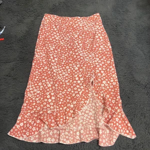 Rosa blommig kjol med slits - Rosa kjol med vitt blommönster och volangslita. Kjolen har en feminin passform och stängs med dragkedja i sidan. Oanvänd utan prislapp.