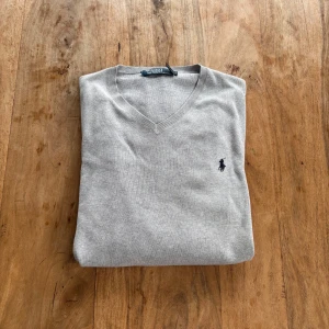 Polo Ralph Lauren – Ljusgrå bomullströja med V-ringning - Mjuk och stilren tröja i finstickad bomull med klassisk V-ringning och marinblå broderad logga.  Storlek: M (normal i storlek) Färg: Ljusgrå med marinblå logga Material: Bomull (etikett ej synlig, men sannolikt 100% bomull) Skick: Mycket fint skick – inga hål eller fläckar Stil: Preppy / Klassisk / Business casual Passform: Smal rak modell med ribbade muddar Nypris: ca 1 200 kr Pris: 300 kr  Frakt: Skickas snabbt & spårbart