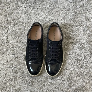 Lanvin cap toe 43-44 - Säljer ett par svarta sneakers från Lanvin med snygg lackad tå och vita sulor. Skorna har snörning och en klassisk rund tå. Perfekta för dig som vill ha en stilren och lyxig look. Skorna kommer med originalpåsar och extra skosnören.