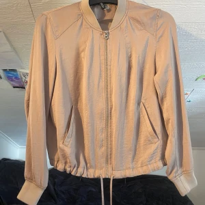 Beige bomberjacka från H&M - Säljer en beige bomberjacka från H&M i storlek M. Jackan har dragkedja framtill, två snedställda fickor och ribbade muddar vid ärmslut och hals. Dragsko i nederkant för justerbar passform. Perfekt för vår och höst.