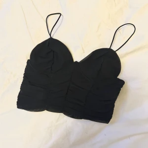 Svart croppad linnetopp med tunna axelband - Säljer en svart croppad linnetopp med tunna axelband och rynkade detaljer framtill. Perfekt för att styla med jeans eller kjol för en trendig look. Använd 1 gång, toppen är som ny👏🏼