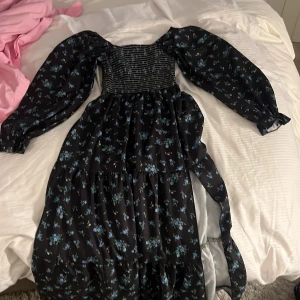 Svart blommig långklänning med puffärmar - Svart långklänning med blått blommönster och smockad överdel. Klänningen har långa puffärmar och ett skärp i midjan. Perfekt för dig som gillar romantisk stil och vill sticka ut lite extra.