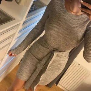 Grå stickad klänning med knytband - Grå stickad klänning med lång ärm och knytband i midjan. Klänningen har en rund halsringning och är i en mjuk, stretchig kvalitet som sitter snyggt på kroppen. Använd 1 gång, från brave soul.