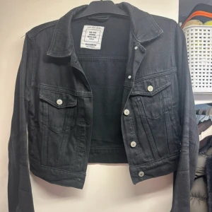 Svart croppad jeansjacka från Pull&Bear - Säljer en svart croppad jeansjacka från Pull&Bear. Jackan har klassisk krage, knappar framtill och två bröstfickor med lock. Perfekt för dig som gillar en trendig och avslappnad stil.