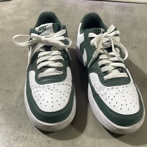Nike Air Force 1 sneakers i vitt och mörkgrönt - Nike Air Force 1 sneakers i vitt och mörkgrönt med klassisk låg siluett. Skorna har snörning, perforerad tå och vit sula. Ikonisk Nike-logga på sidan och bak. Perfekta för en sportig och stilren look.