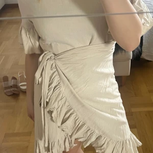 Beige omlottklänning med volang - Supersöt beige omlottklänning med volangdetaljer och knyt i midjan. Klänningen har korta ärmar med lätt puff och är tillverkad i ett tunt, luftigt material. Perfekt för varma dagar när du vill känna dig både söt och bekväm.