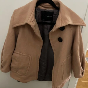 Rosa kappa - Säljer en rosa kort kappa från Zara Basic med stora svarta knappar framtill och bred krage. Jackan har två fickor och en snygg detalj med knappar i ryggen. Perfekt för dig som vill ha en stilren och trendig look.