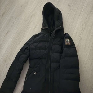 Svart dunjacka med huva från Parajumpers - Säljer en svart dunjacka från Parajumpers med huva och dragkedja framtill. Jackan har två snedställda fickor med dragkedja och en patch på ena ärmen. Perfekt för kyliga dagar.