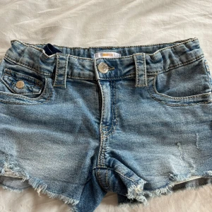 Blå jeansshorts från Gina Tricot - Säljer ett par klassiska blå jeansshorts från Gina Tricot. Shortsen har fransade benslut, fem fickor och silverfärgade knappar. Perfekta för sommaren ! har aldrig använts, inga fläckar eller hål.
