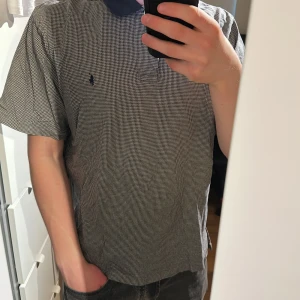 Grå- och vitmönstrad pikétröja från Ralph Lauren - Säljer en klassisk pikétröja från Ralph Lauren i grått och vitt småmönster med mörkblå krage och knappslå. Liten broderad logga på bröstet. Perfekt för en stilren och avslappnad look. Gamla taggen 