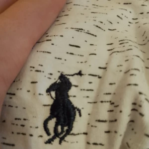 Vit t-shirt med svart mönster från Polo Ralph Lauren - Säljer en vit t-shirt från Polo Ralph Lauren med diskret svart mönster och klassisk broderad logga på bröstet. Skön passform och perfekt för en avslappnad stil. Materialet känns mjukt och andas bra.