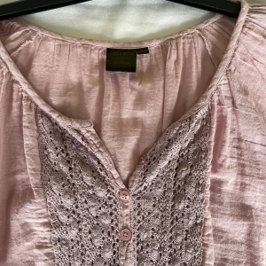 Ljusrosa blus med spetsdetaljer från Cellbes - Säljer en ljusrosa blus från Cellbes med vackra spetsdetaljer och knappar framtill. Blusen har långa ärmar och är tillverkad i ett lätt, glansigt material. Perfekt för dig som gillar romantisk stil.