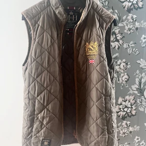 Olivgrön quiltad väst från Morris - Säljer en olivgrön quiltad väst från Morris med broderad guldfärgad logga och brittisk flagga på bröstet. Västen har ståkrage och dragkedja framtill. Perfekt för lager-på-lager och snygg detalj till din outfit. Hör av er vid minsta fråga!
