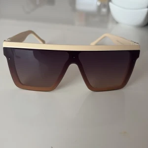 Beige oversized solglasögon med guldiga detaljer - Snygga oversized solglasögon i beige med markerad guldig ram upptill och mörka, fyrkantiga linser. Sidorna har coola metalldetaljer i guld med nitar. Perfekt accessoar för en trendig look.