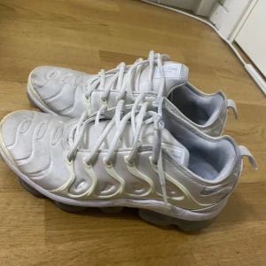 Vita Nike Air Vapormax Plus sneakers - Säljer ett par vita Nike Air Vapormax Plus sneakers med tydlig logga bak och på plösen. Skorna har en futuristisk design med synliga luftbubblor i sulan och snörning framtill. Perfekta för dig som gillar streetwear och sportig stil.
