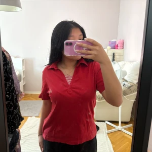 Röd pikétröja  - Säljer en klassisk röd pikétröja med korta ärmar och krage. Tröjan har en liten broderad logga med ankare på bröstet och en randig detalj i halsringningen. Perfekt för en avslappnad och sportig stil eller vintage