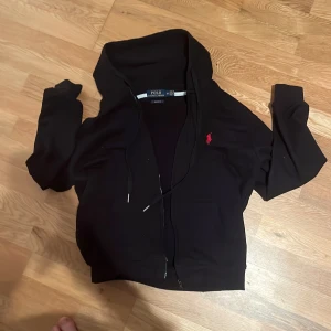 Svart hoodie från Polo Ralph Lauren - Svart hoodie från Polo Ralph Lauren med dragkedja och huva. Klassisk röd broderad logga på bröstet och fickor framtill. Modellen är i custom fit och har ribbade muddar vid ärmslut och nederkant. Perfekt för en avslappnad stil.